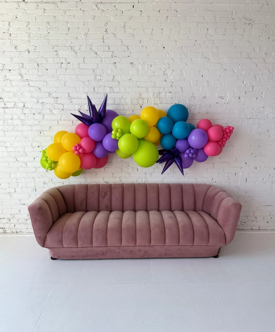 Large Party-Ready Balloon Garland with Foil Starburst Balloons by Lady With The Balloons in Leawood, Kansas — bright, colorful balloon garland displayed above a mauve sofa against a white brick wall, featuring bold foil accents for added shine and dimension.