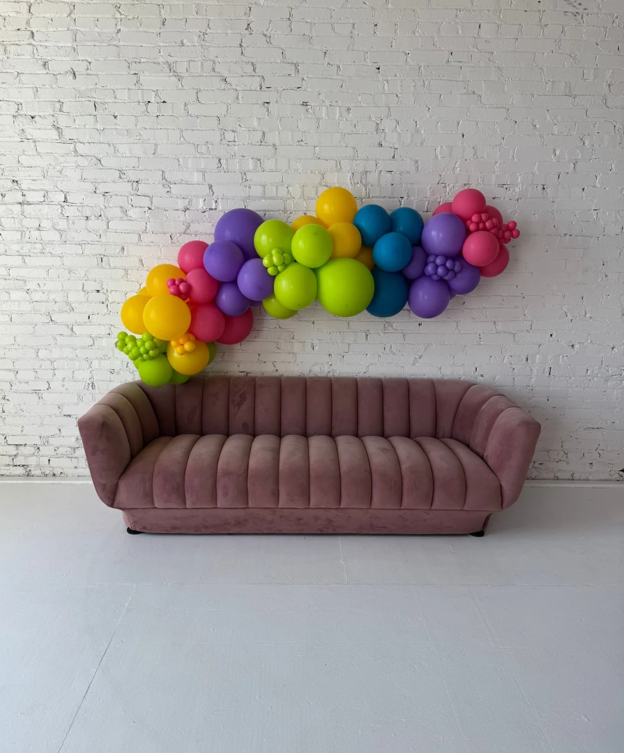 Large Party-Ready Balloon Garland by Lady With The Balloons in Leawood, Kansas — bright, colorful balloon garland displayed above a mauve sofa against a white brick wall, designed without foil balloons for a smooth, modern finish