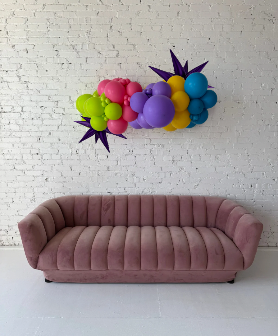 Party-Ready Grab & Go Balloon Garland by Lady With The Balloons in Leawood, Kansas — bright, colorful balloon garland with starburst accents displayed above a mauve sofa against a white brick wall.
